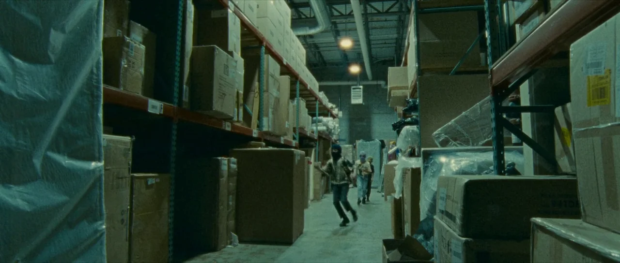 Riddle of Fire (2023) movie still: wide — A long, narrow aisle in a large warehouse is filled with stacked cardboard shipping boxes. Two individuals…