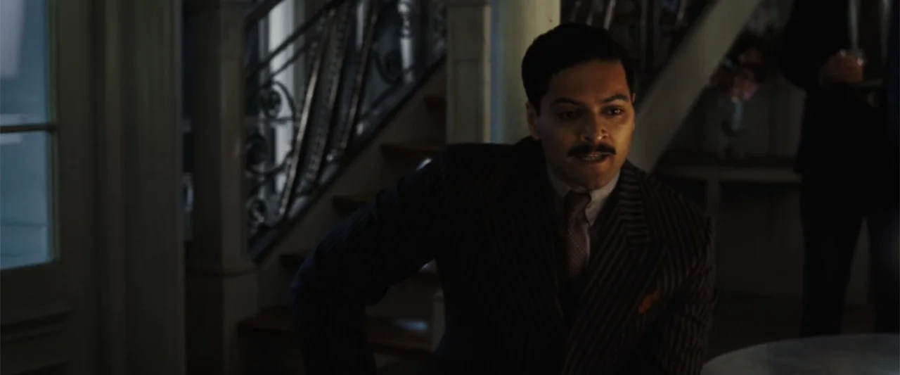 Death on the Nile (2022) movie still: medium — A dark room features a wooden staircase with an ornate metal railing in the background. A man in a…