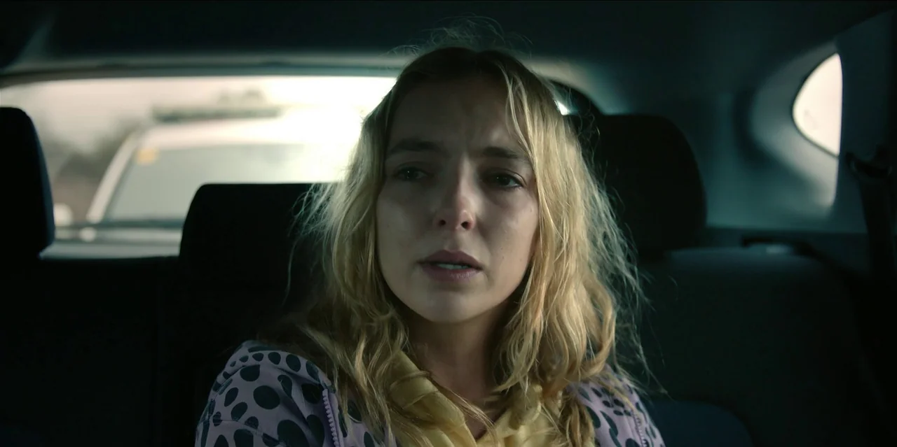 Help (2021) movie still: closeup — The interior of a vehicle shows a woman sitting in the back seat. She looks forward while light from the window…