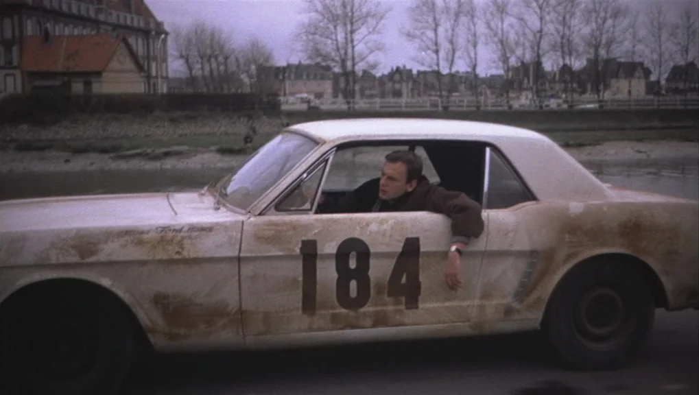 Un Homme et Une Femme (1966) movie still: wide — A dirty, light-colored Ford Mustang with the number 184 painted on its side drives. The car passes…