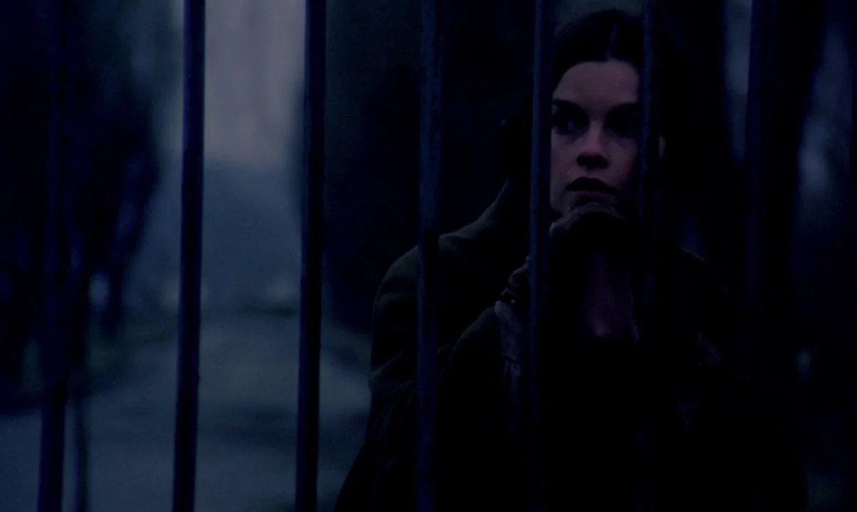 Kamouraska (1973) movie still: closeup — An iron railing partially obscures a young woman standing at night. She gazes out into the dim space while…