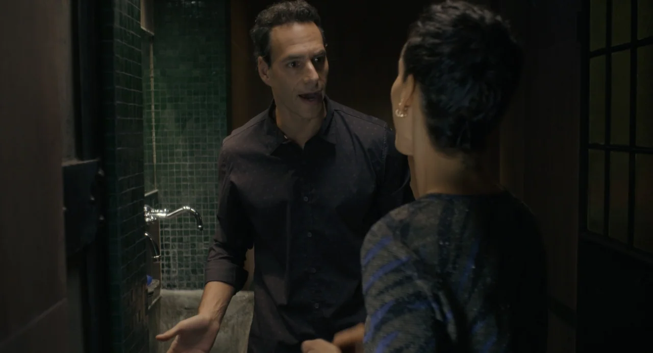 Little White Lies (2010) movie still: medium — A dimly lit restroom features dark green mosaic tiles and a chrome faucet fixture. A man gestures…
