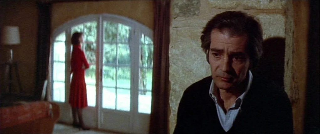 Love Unto Death (1984) movie still: medium — A rustic interior features stone walls and large arched glass doors leading to greenery. A man sits in…
