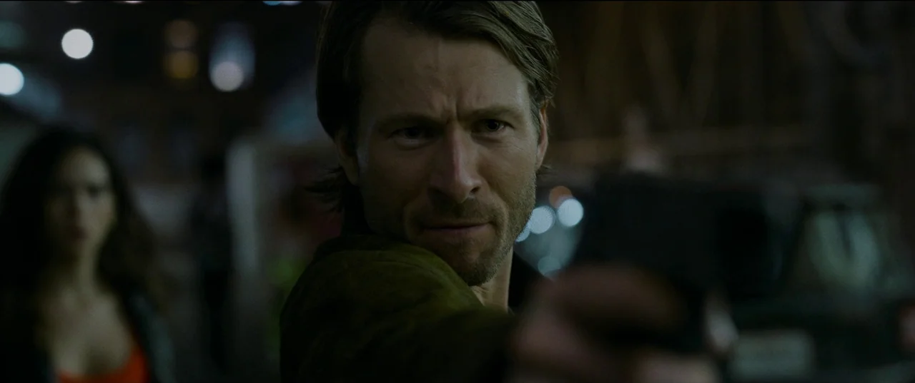 Hit Man (2023) movie still: closeup — An interior space features low-light conditions with blurred bokeh lights in the distance. A man in the…