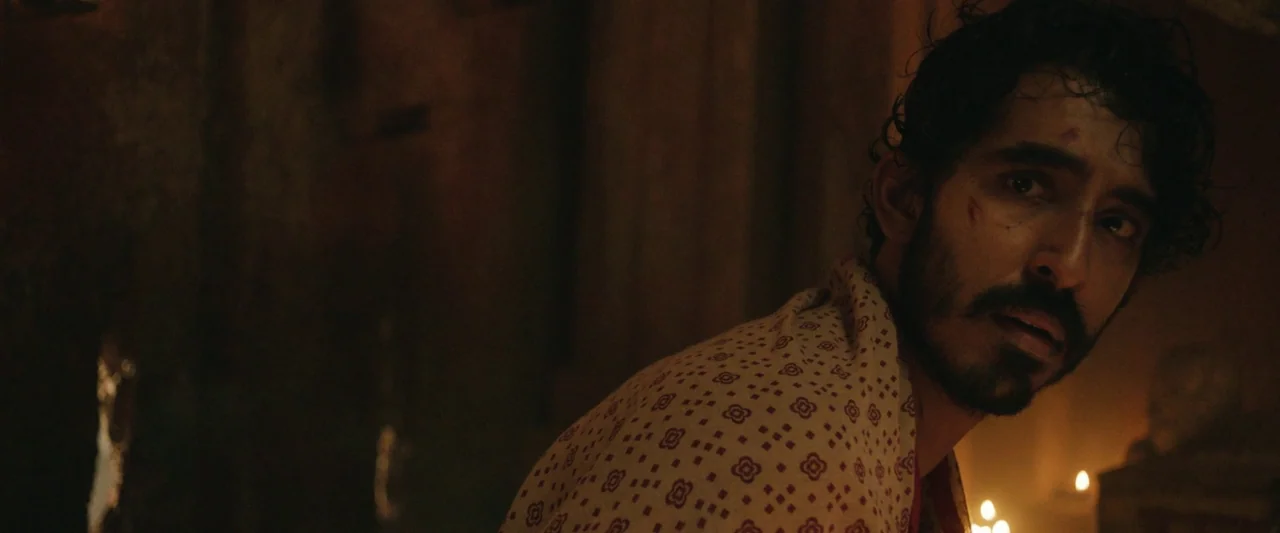 Monkey Man (2024) movie still: closeup — A dimly lit interior space with dark walls and soft candlelight illuminating the frame. A man with a beard…