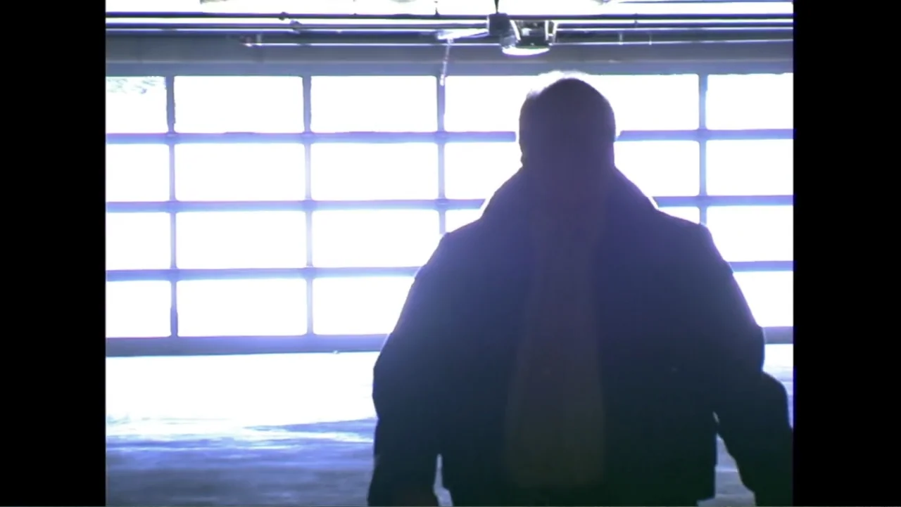 Savageland (2015) movie still: medium — A dark, enclosed space features a large window with horizontal bars and bright daylight. A man in a dark…