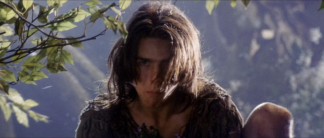 Legend (1985) movie still: closeup — Dense foliage frames a man within a sunlit, shadowed wooded environment. He looks directly at the viewer with…