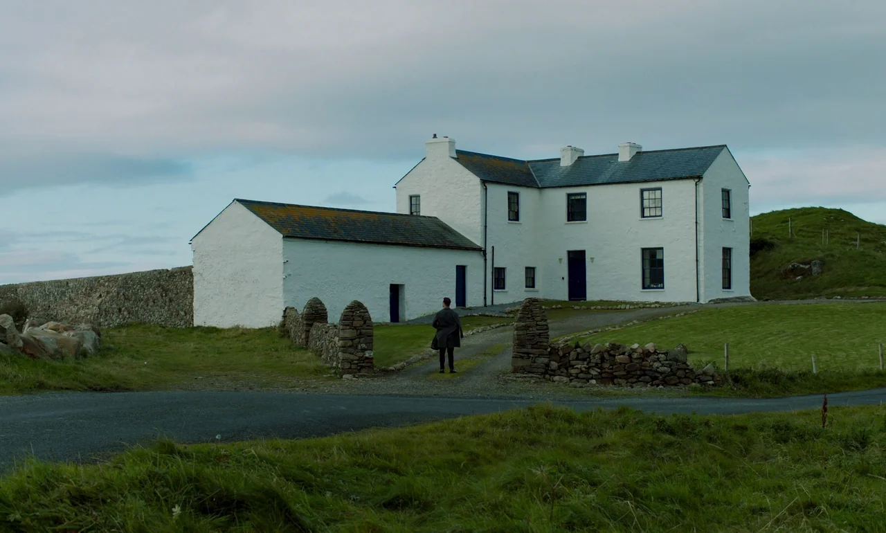 It Is In Us All (2022) movie still: wide — A white two-story farmhouse with a stone wall sits in a grassy field. A man in a dark coat walks toward…