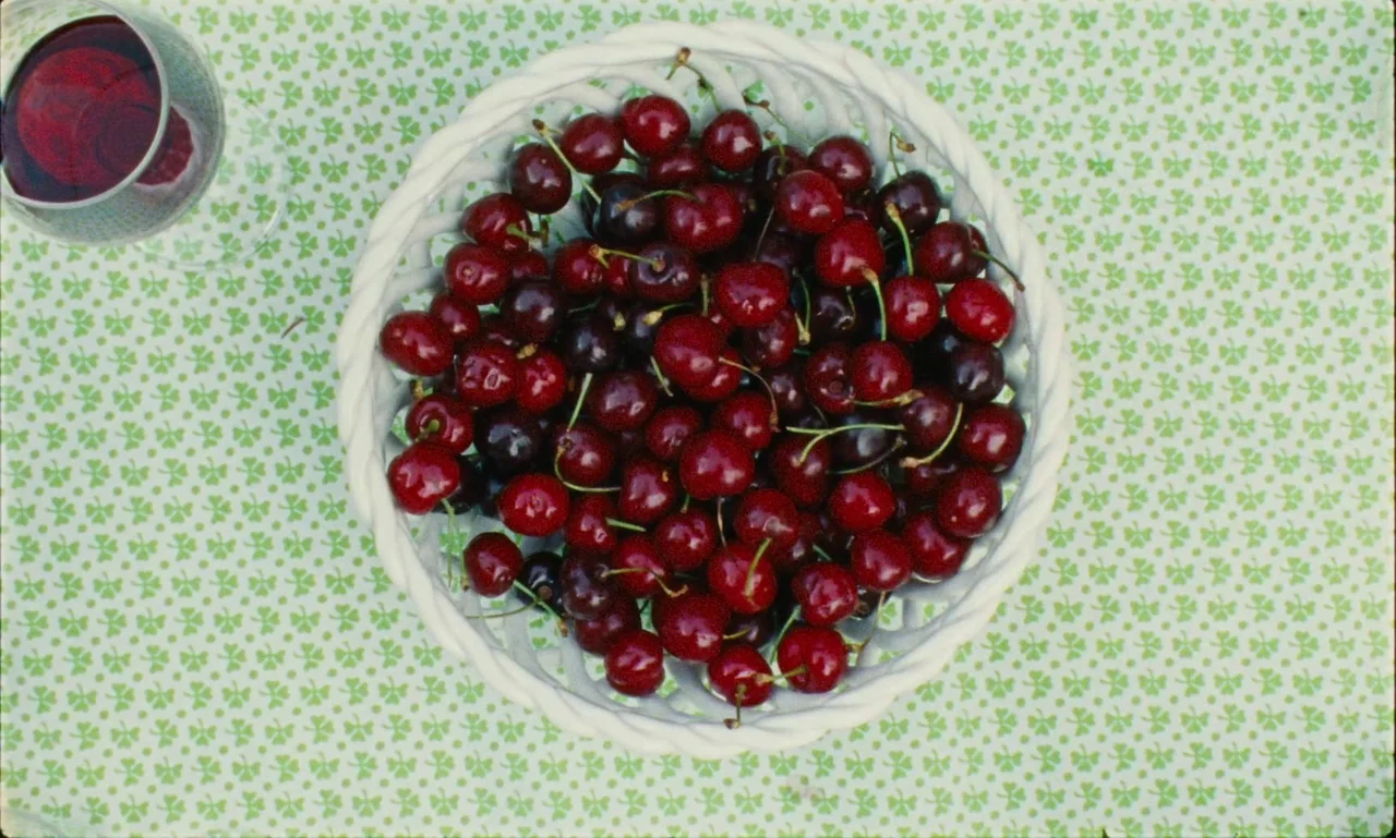 Just The Two Of Us (2023) movie still: closeup — A white woven basket filled with dark red cherries rests on a shamrock-patterned tablecloth. A…