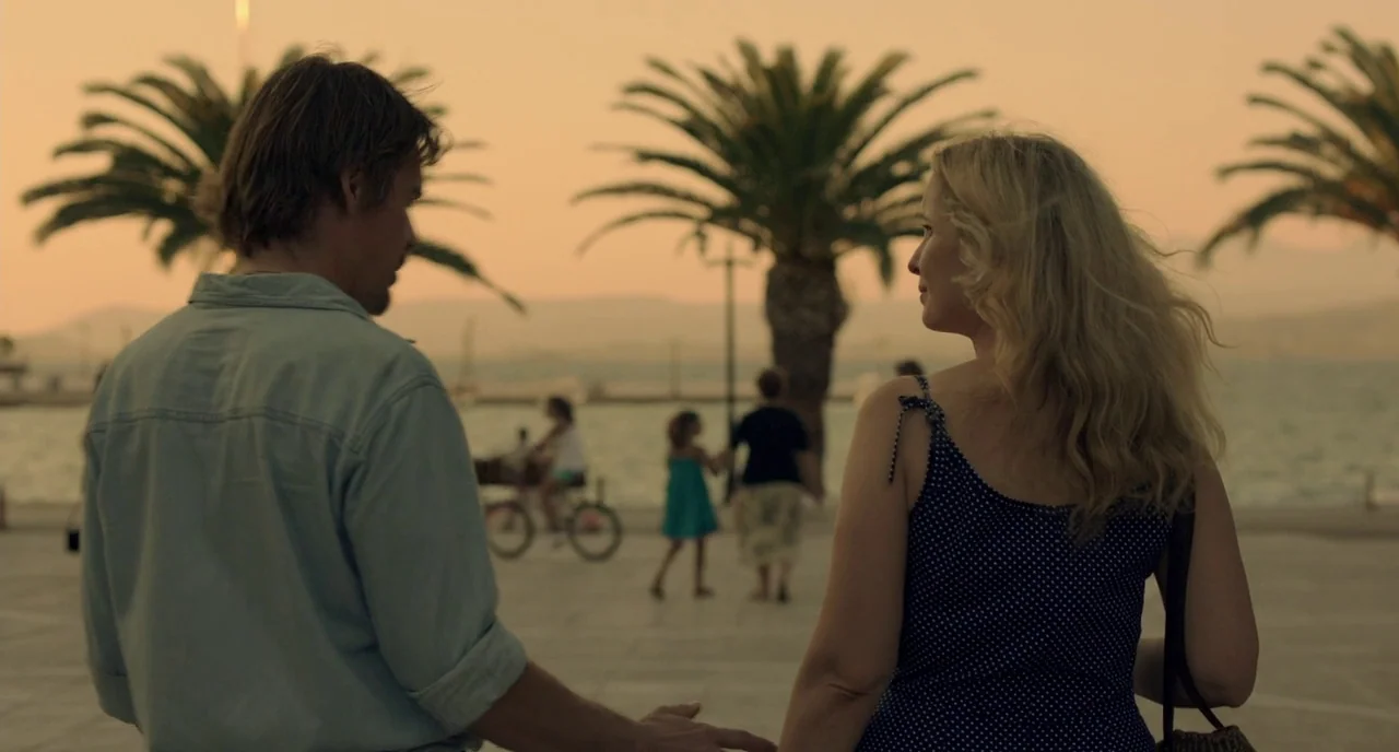 Before Midnight (2013) movie still: medium — A stone-paved waterfront promenade features palm trees and a view of the sea. A man and a woman walk…