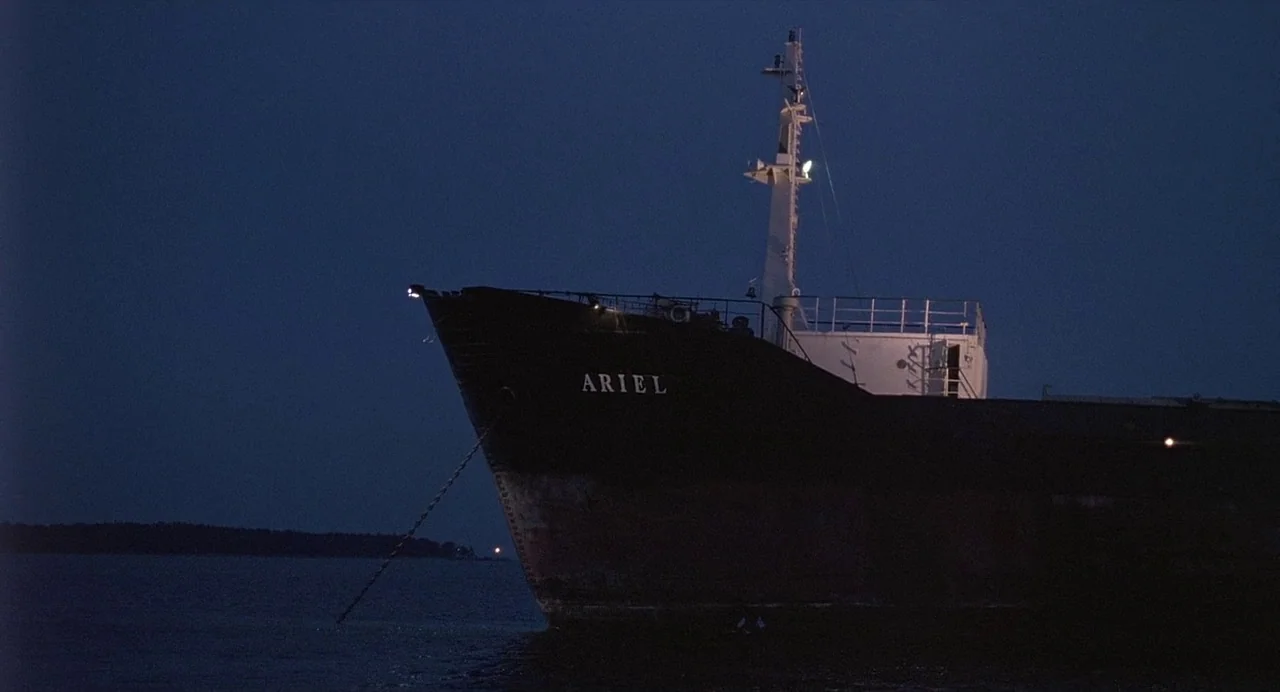 Ariel (1988) movie still: wide — A large cargo ship with the name Ariel painted on its bow sits at night. The vessel is anchored in dark water under…