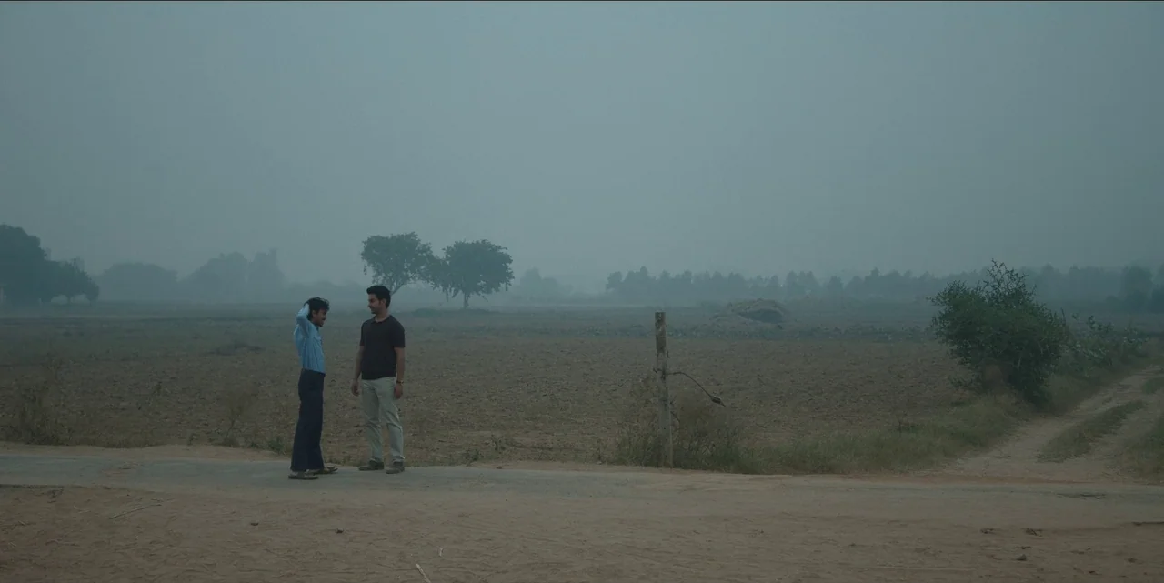 The White Tiger (2021) movie still: wide — A flat rural landscape stretches toward a distant tree line under hazy conditions. Two men stand on a…