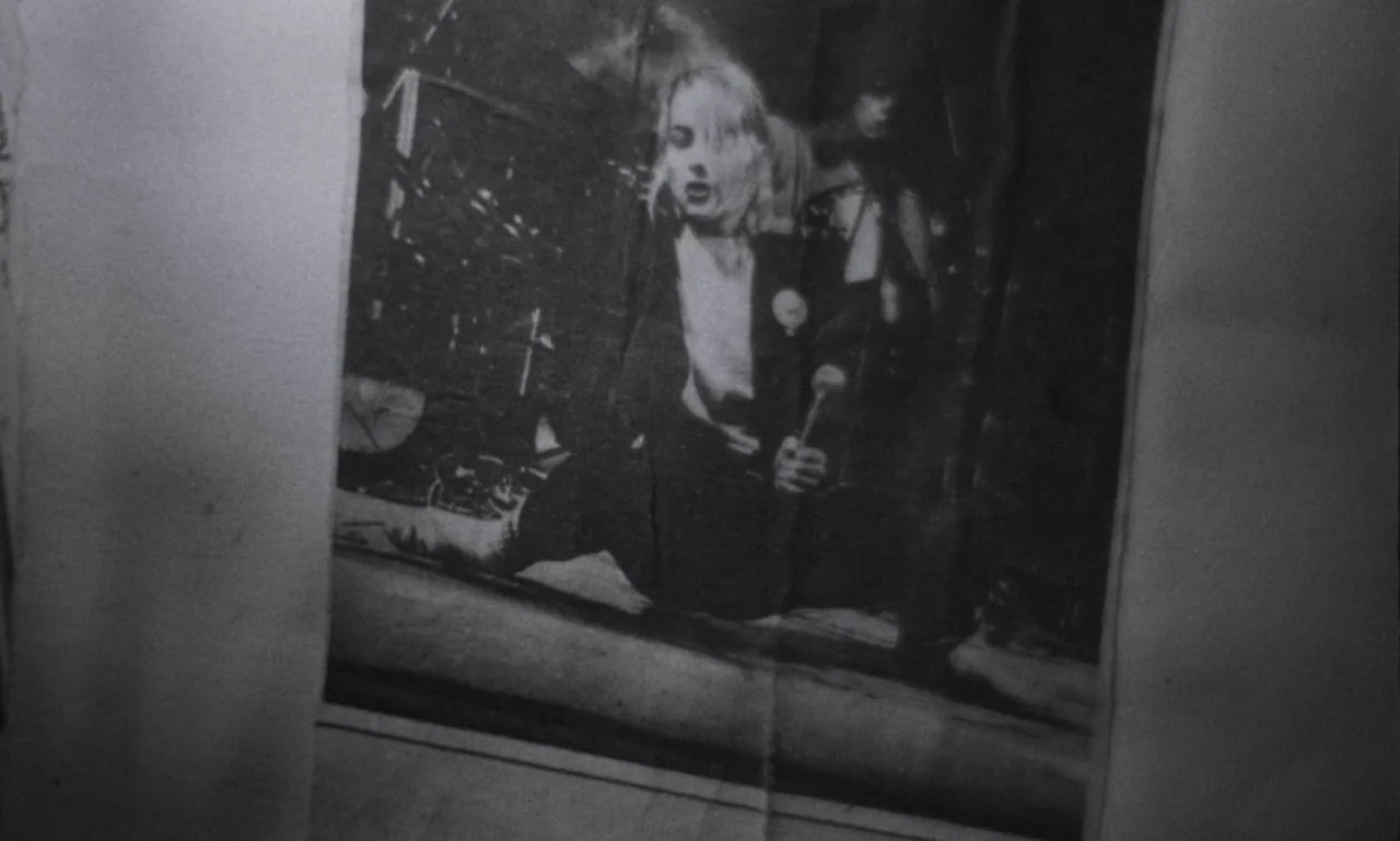 The Secret Son (1979) movie still: closeup — A grainy black and white newspaper print shows a photograph of a performer. A woman with blonde hair…