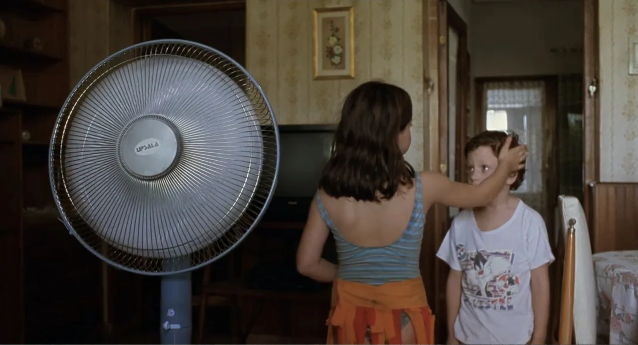 La Ciénaga (2001) movie still: medium — A living room features floral patterned wallpaper and an oscillating pedestal fan in foreground. A young…