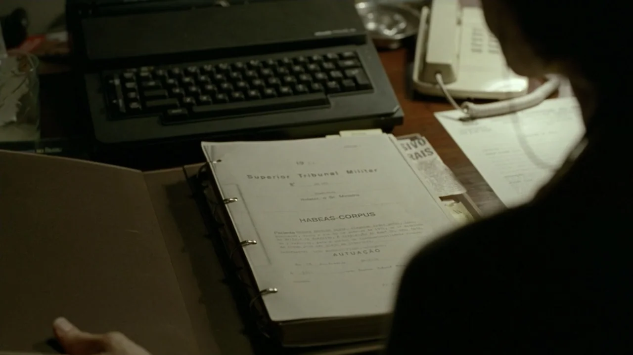 I'm Still Here (2024) movie still: medium — A dimly lit office desk contains a typewriter, a landline telephone, and a legal file. A person views a…