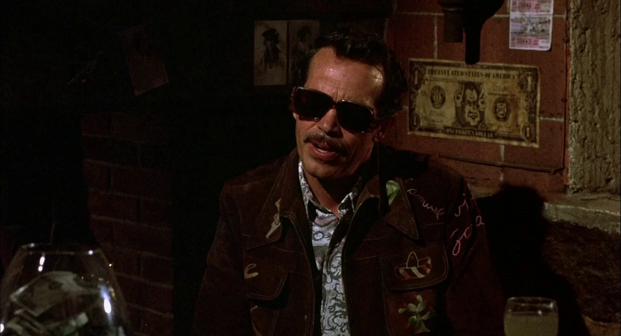 Bring Me The Head of Alfredo Garcia (1974) movie still: medium — A dimly lit room features a brick wall adorned with novelty paper currency. A man…