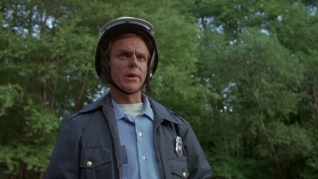 Friday the 13th (1980) movie still: medium — A police officer stands in a dense wooded area with blurred green trees behind. He wears a motorcycle…