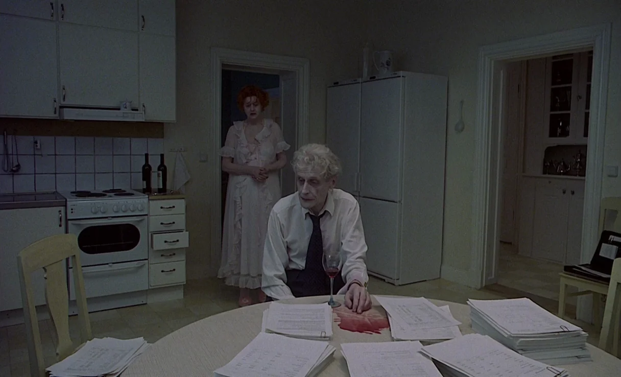 Songs from the Second Floor (2000) movie still: medium — A sparse, functional home dining area with white cabinetry and multiple stacks of…