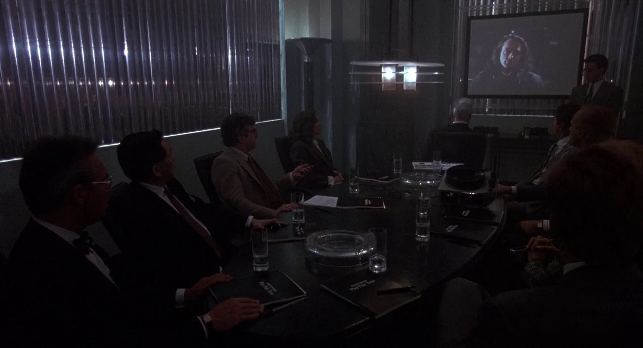 Child’s Play 3 (1991) movie still: wide — A dark boardroom features a large oval table surrounded by people in business attire. A video of a person…