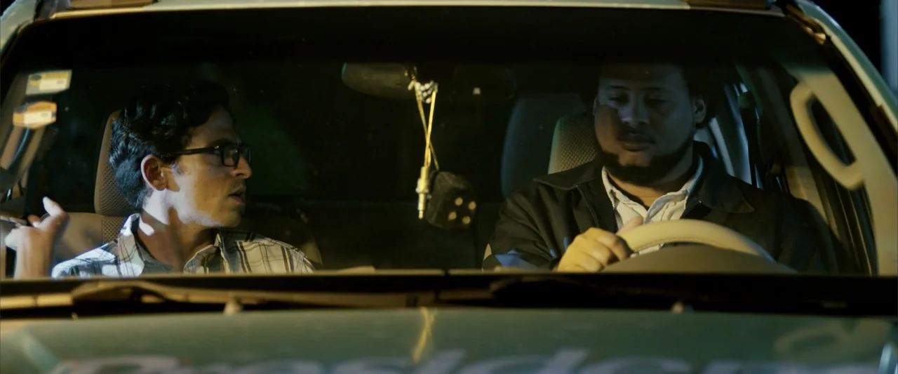 La Musiquita por Dentro (2019) movie still: medium — A nighttime view inside a car cabin with two men sitting in the front. The driver steers while…