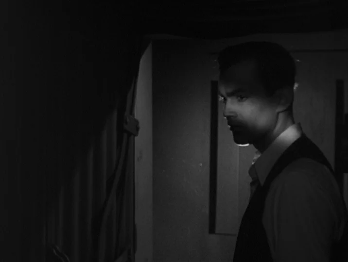 Thirst (1949) movie still: medium — A dimly lit room with heavy shadows obscuring much of the background environment. A man in a vest stands looking…