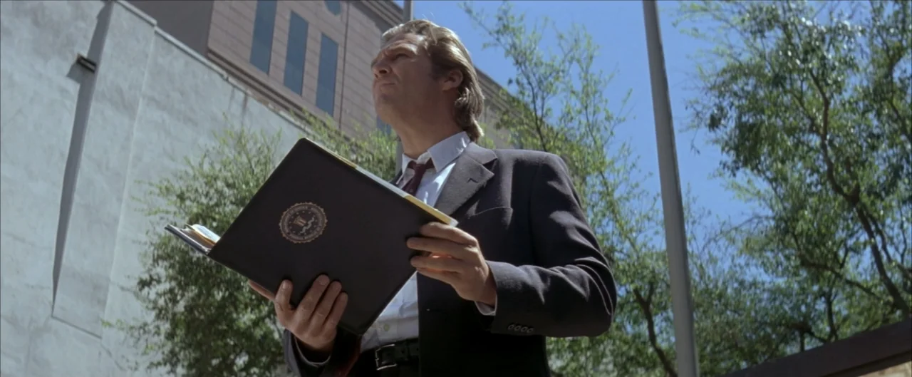 Arlington Road (1999) movie still: medium — A concrete building wall stands behind a man in a business suit. The man holds an open black binder with…