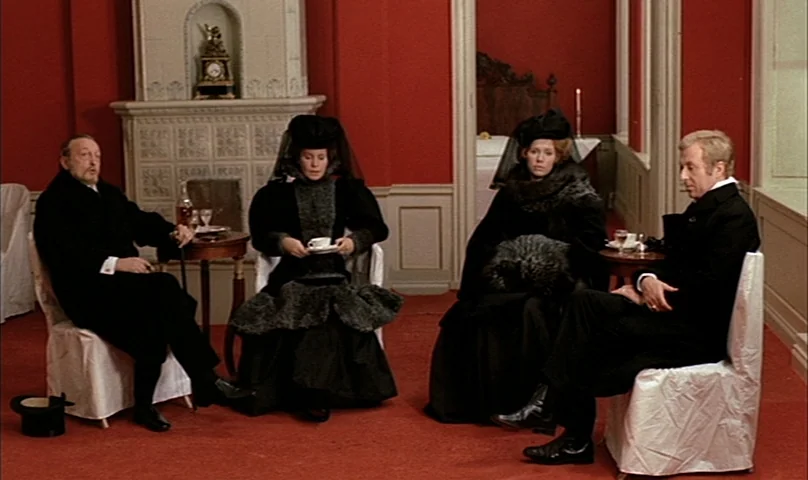 Cries and Whispers (1972) movie still: wide — A spacious room with bright red walls and white wainscoting features a tiled fireplace. Four people in…