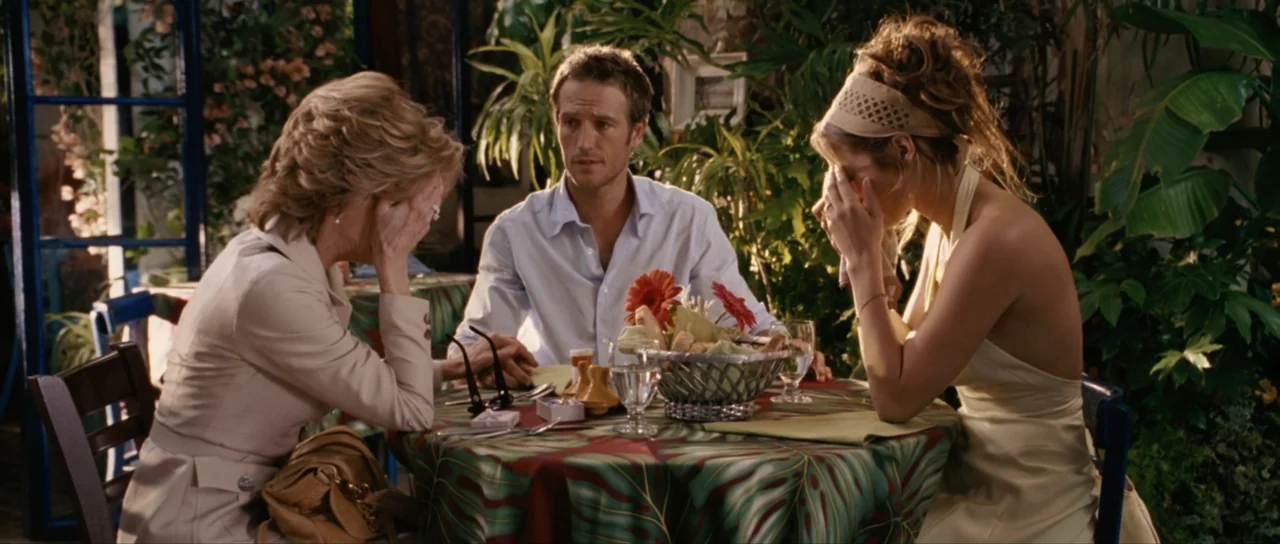 Monster-in-Law (2005) movie still: medium — A patio dining area surrounded by lush green tropical plants and dark wood structures. Two women and one…