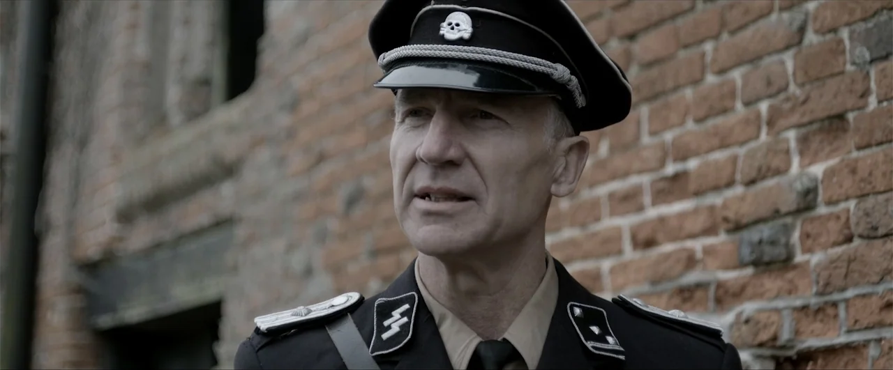 Behind the Line: Escape to Dunkirk (2020) movie still: medium — A man in a German officer uniform stands before a decaying weathered brick wall. The…