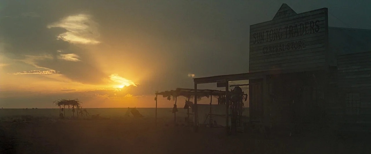 The Proposition (2005) movie still: establishing — An isolated wooden trading post stands on a vast, flat desert landscape at sunset. Sun rays cut…