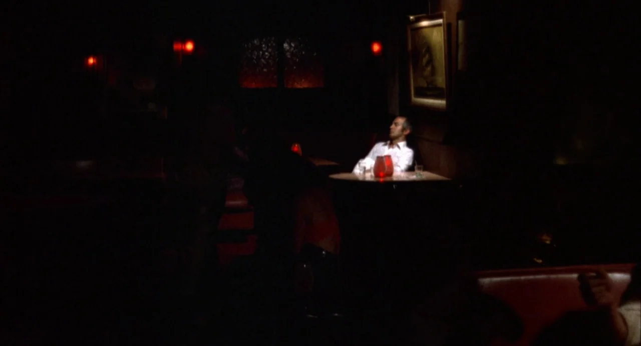 The Killing of a Chinese Bookie (1976) movie still: wide — A dark, dimly lit bar with red leather booths and a small round table. A man in a white…