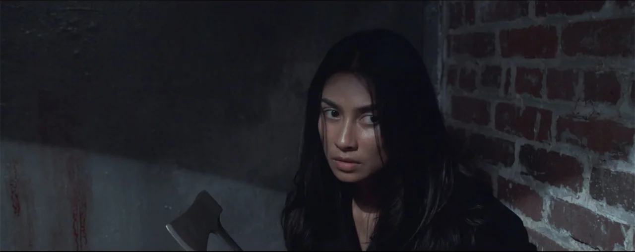 El Último Juego (2025) movie still: medium — A dark corner of a basement with an exposed red brick wall and concrete floor. A woman with long dark…
