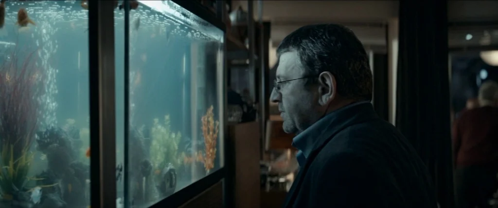 Graduation (2016) movie still: medium — A dimly lit room features a large rectangular aquarium filled with artificial plants. A middle-aged man in a…