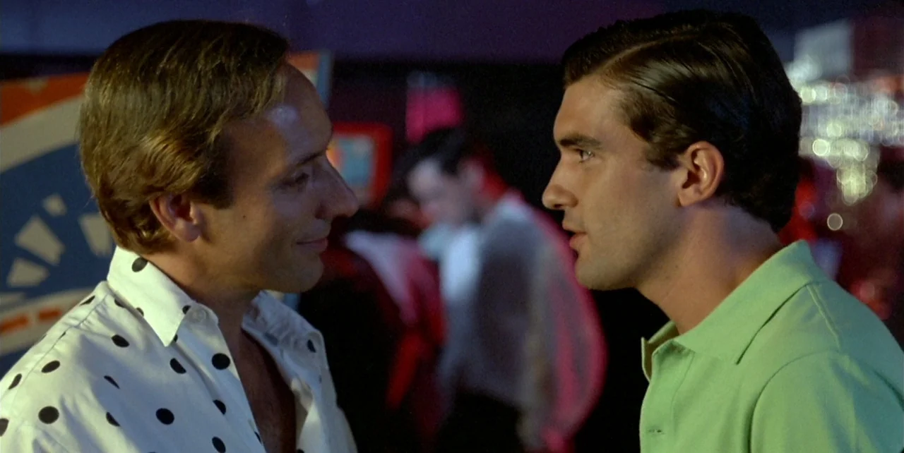 Law of Desire (1987) movie still: medium — A dimly lit nightclub features blurry silhouettes in the background behind two speaking men. One man…