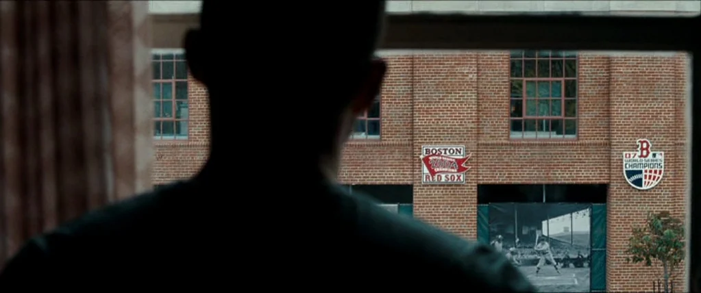 The Town (2010) movie still: medium — A view through a window shows a brick building wall with sports championship banners. A person in the…