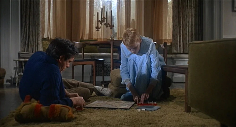 Rosemary’s Baby (1968) movie still: medium — A 1960s living room features a shag carpet and sheer curtains over a large window. A man and a woman…