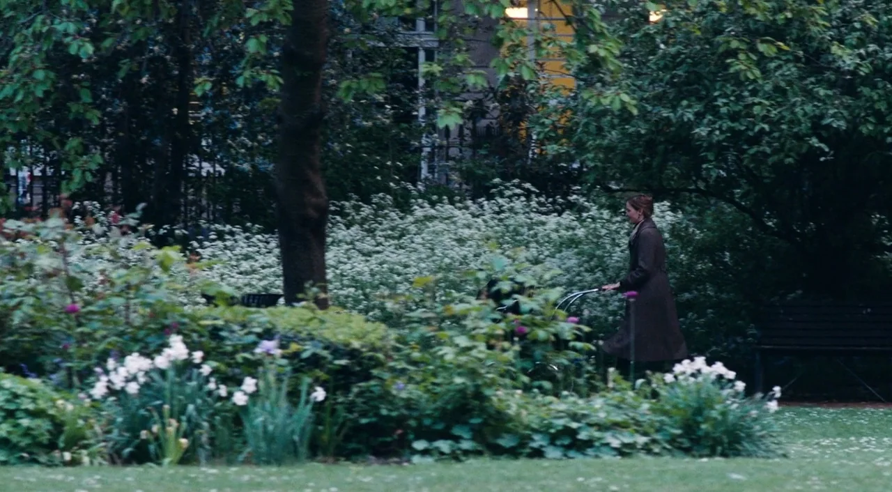Phantom Thread (2017) movie still: wide — A dense park landscape filled with flowering shrubs and large mature trees. A woman in a dark coat pushes…