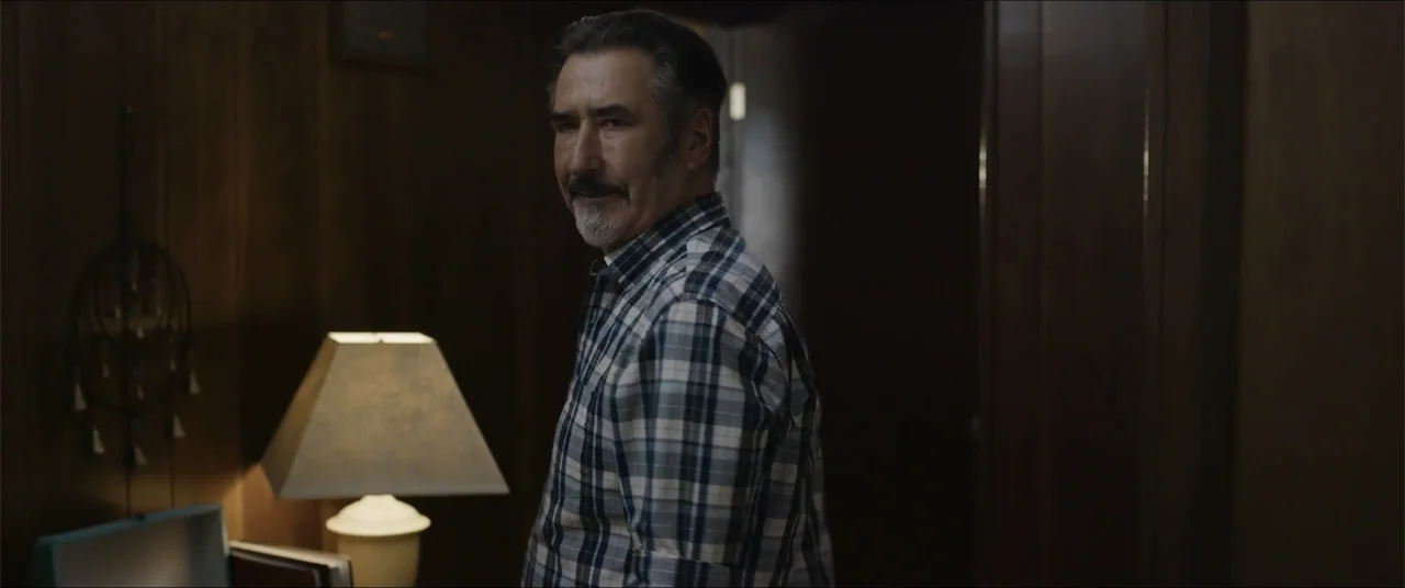 Captive (2020) movie still: medium — A dimly lit room features wood-paneled walls and a lamp casting soft light. A middle-aged man in a plaid shirt…