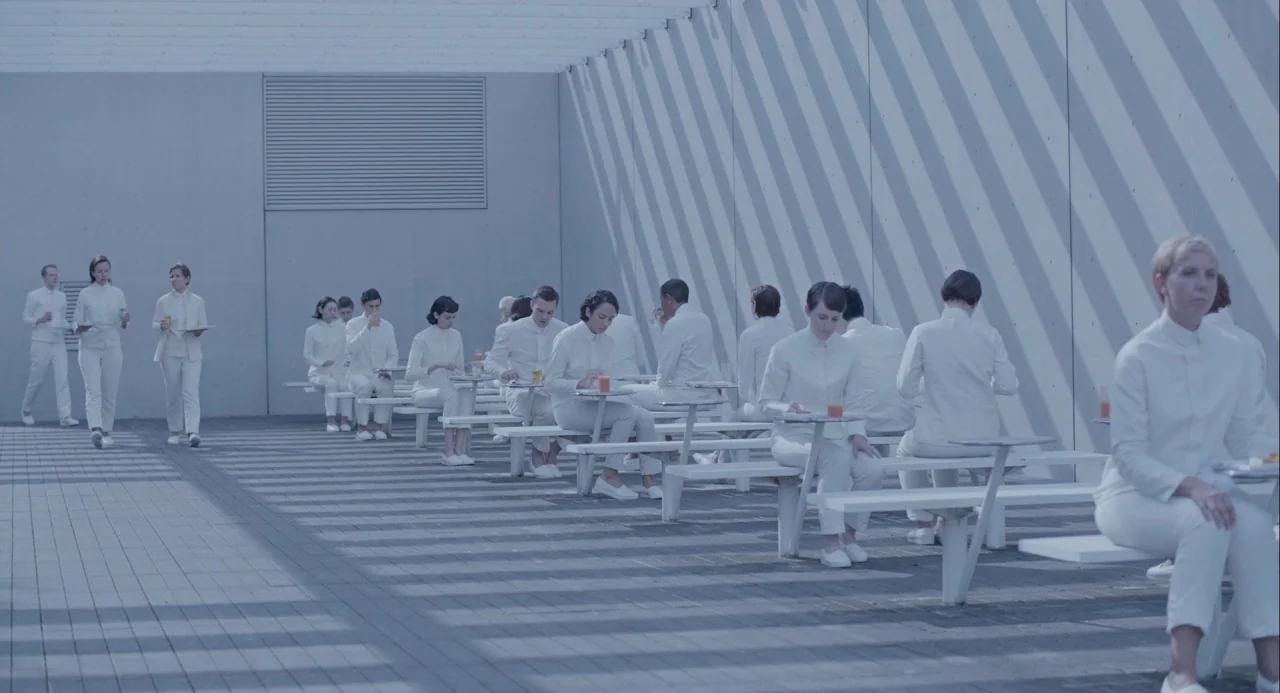 Equals (2015) movie still: wide — A minimalist, open-air concrete dining area features rows of uniform tables under a pergola. Many individuals…