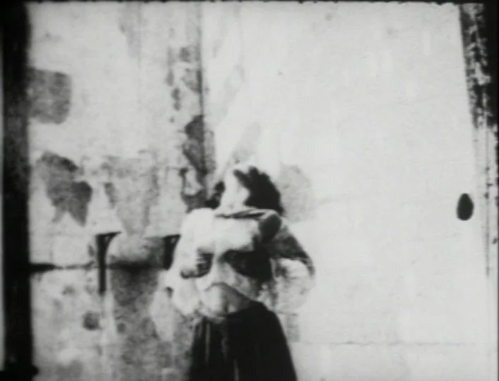 Begotten (1989) movie still: fullbody — A person stands in the corner of a room with weathered, stained walls. The subject is positioned facing…