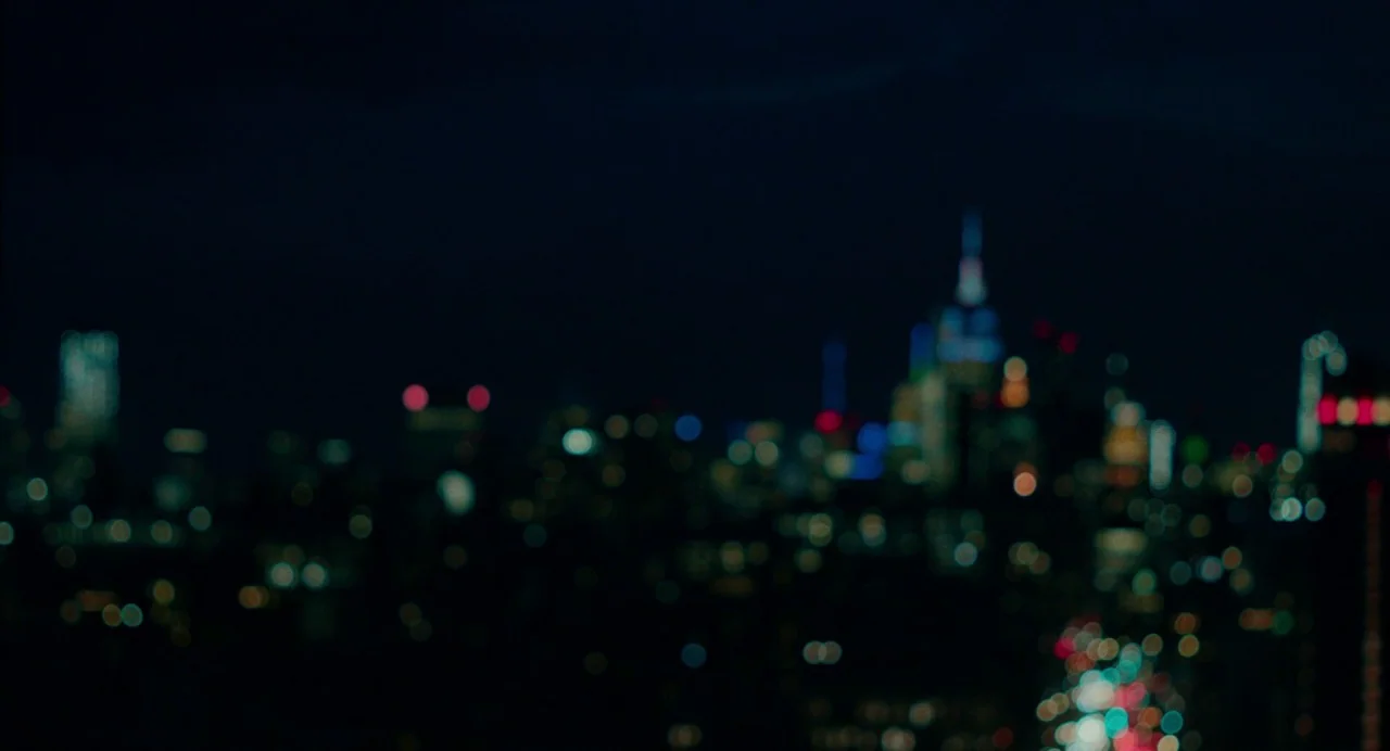 On The Rocks (2020) movie still: establishing — An out-of-focus nocturnal urban skyline glows against a dark, featureless night sky. Clusters of…