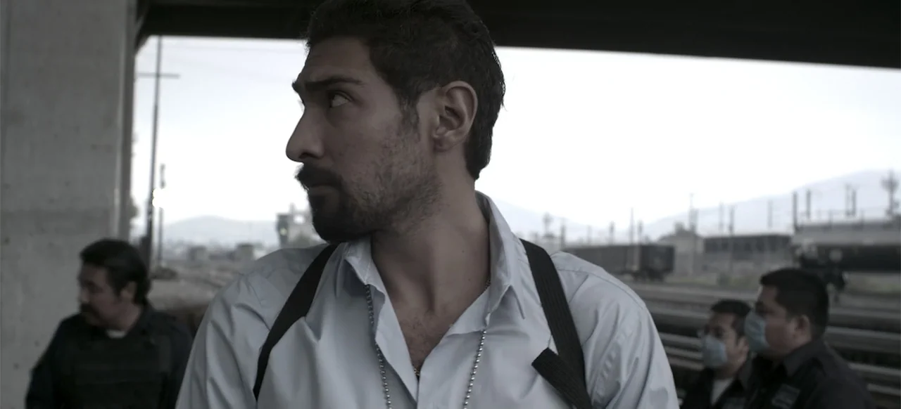 About the Deaths (2016) movie still: medium — A man in a white shirt stands under an industrial concrete bridge by tracks. He wears a shoulder…