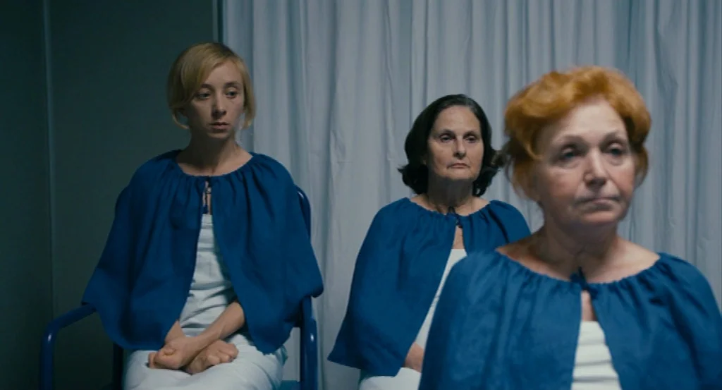 Lourdes movie still: medium — A white curtain hangs behind three women seated in blue medical-style chairs. They all wear identical blue capes over…