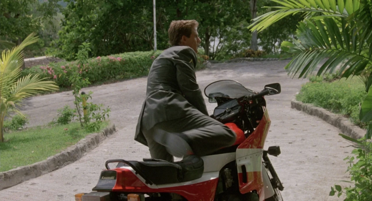 American Ninja (1985) movie still: medium — A paved path leads through lush greenery on a bright sunny day. A man in a suit mounts a red and white…