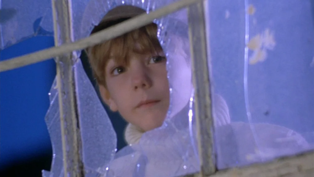 War Requiem (1989) movie still: closeup — A young boy observes the outside world through the shattered panes of a window. He wears a knit sweater…