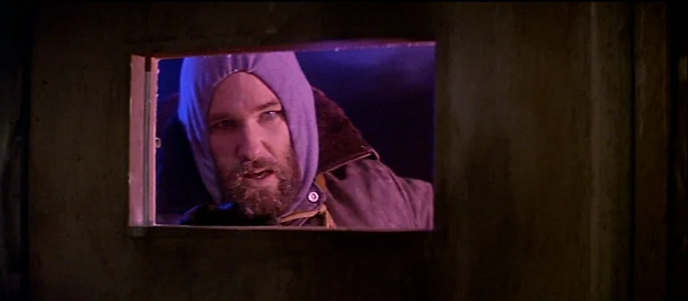 The Thing (1982) movie still: closeup — A man peers through a small rectangular viewport in a dark wooden door. The surrounding area is bathed in a…