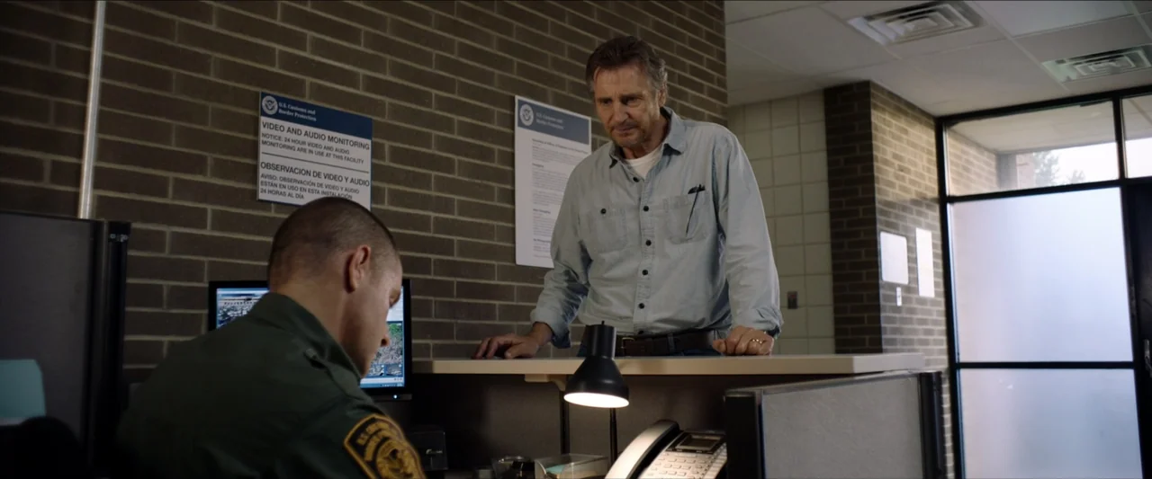 The Marksman (2021) movie still: medium — A government facility features tan brick walls with posted regulatory signage and office cubicle…