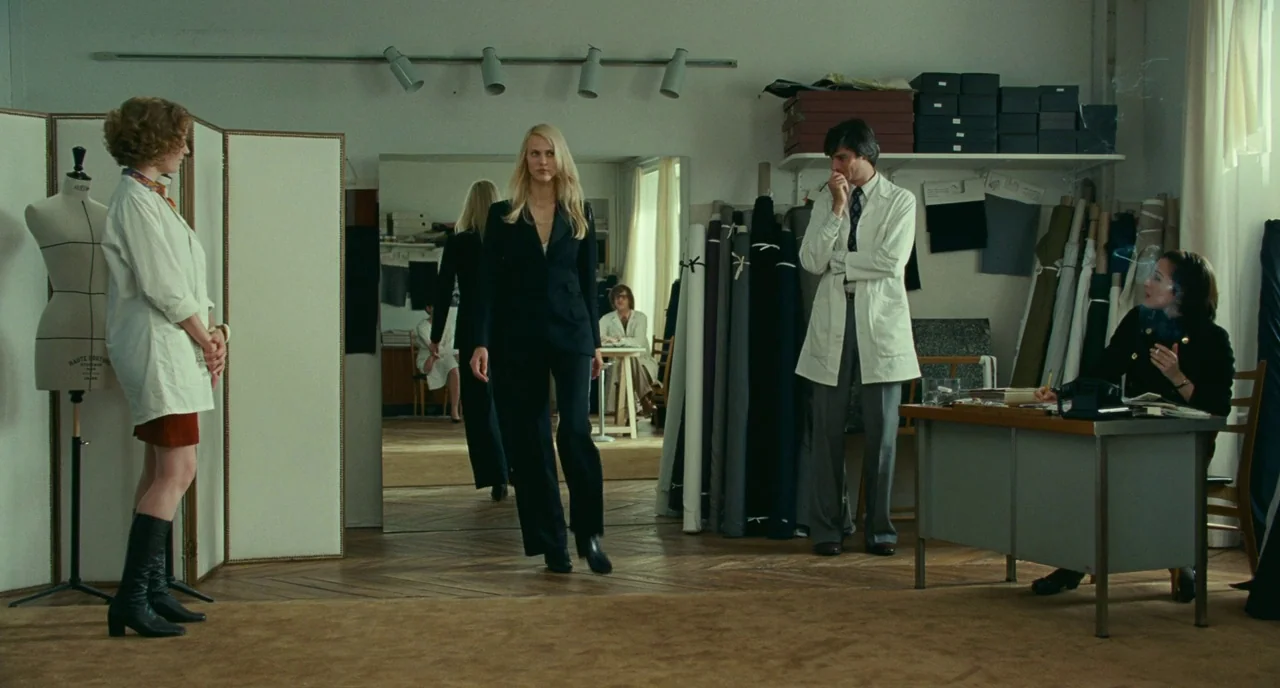 Saint Laurent (2014) movie still: wide — A spacious fashion workroom contains rolls of fabric, a desk, a mirror, and mannequins. A blonde model…