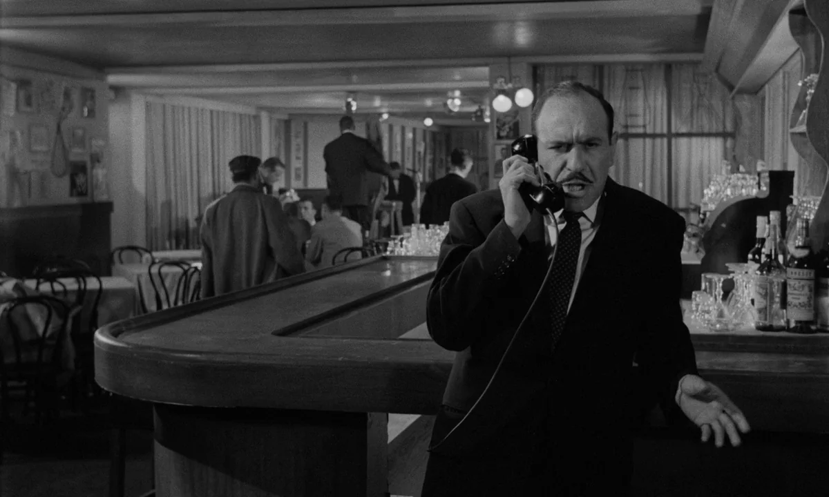 Sweet Smell of Success (1957) movie still: medium — A dimly lit bar with a wooden counter and tables in the background. A man in a suit stands by…