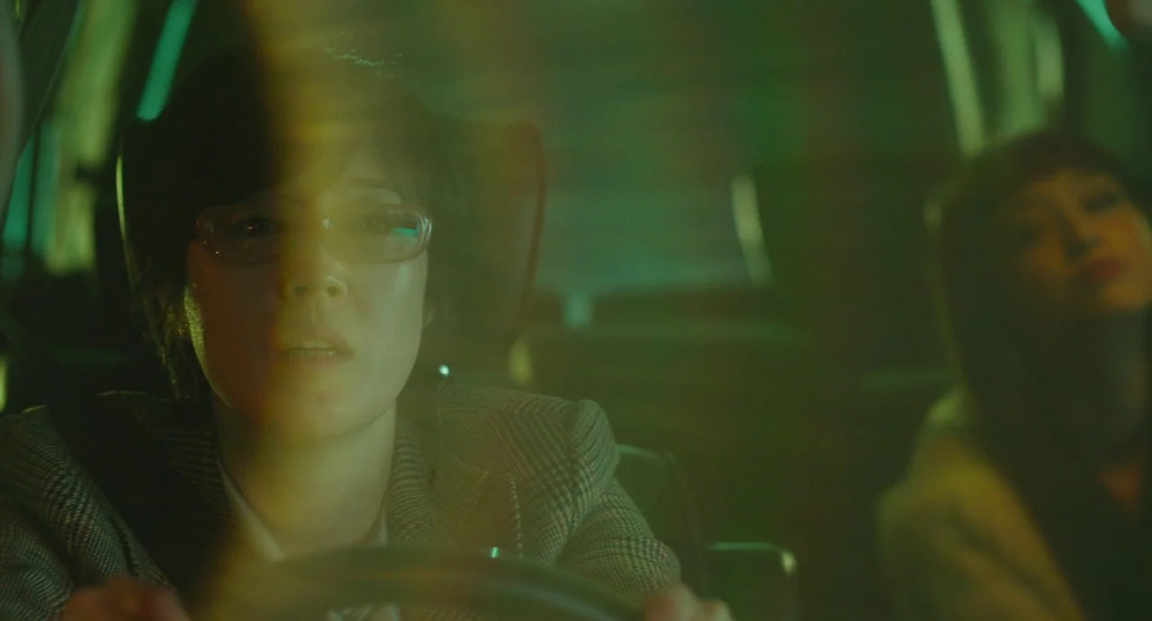 Helter Skelter (2012) movie still: medium — The interior of a moving vehicle is bathed in a distinct green light at night. A woman drives while…